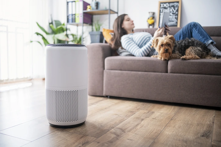 A large, modern, white cylindrical air purifier with a perforated base stands on a wooden floor in the foreground. In the blurred background, a woman is relaxing on a couch with a small dog, using a tablet.
