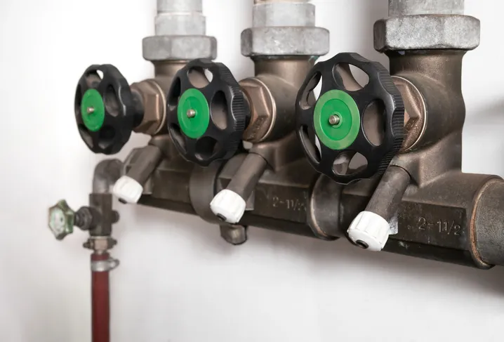 A close-up view of a backflow prevention device featuring a row of three green and black valves, and smaller white-capped valves, attached to a large pipe system against a white wall.