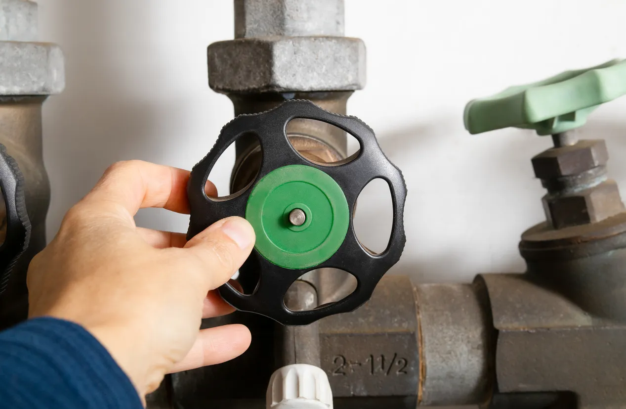 A close-up shot of a hand turning the green and black handle on a backflow preventer valve on a large water pipe.