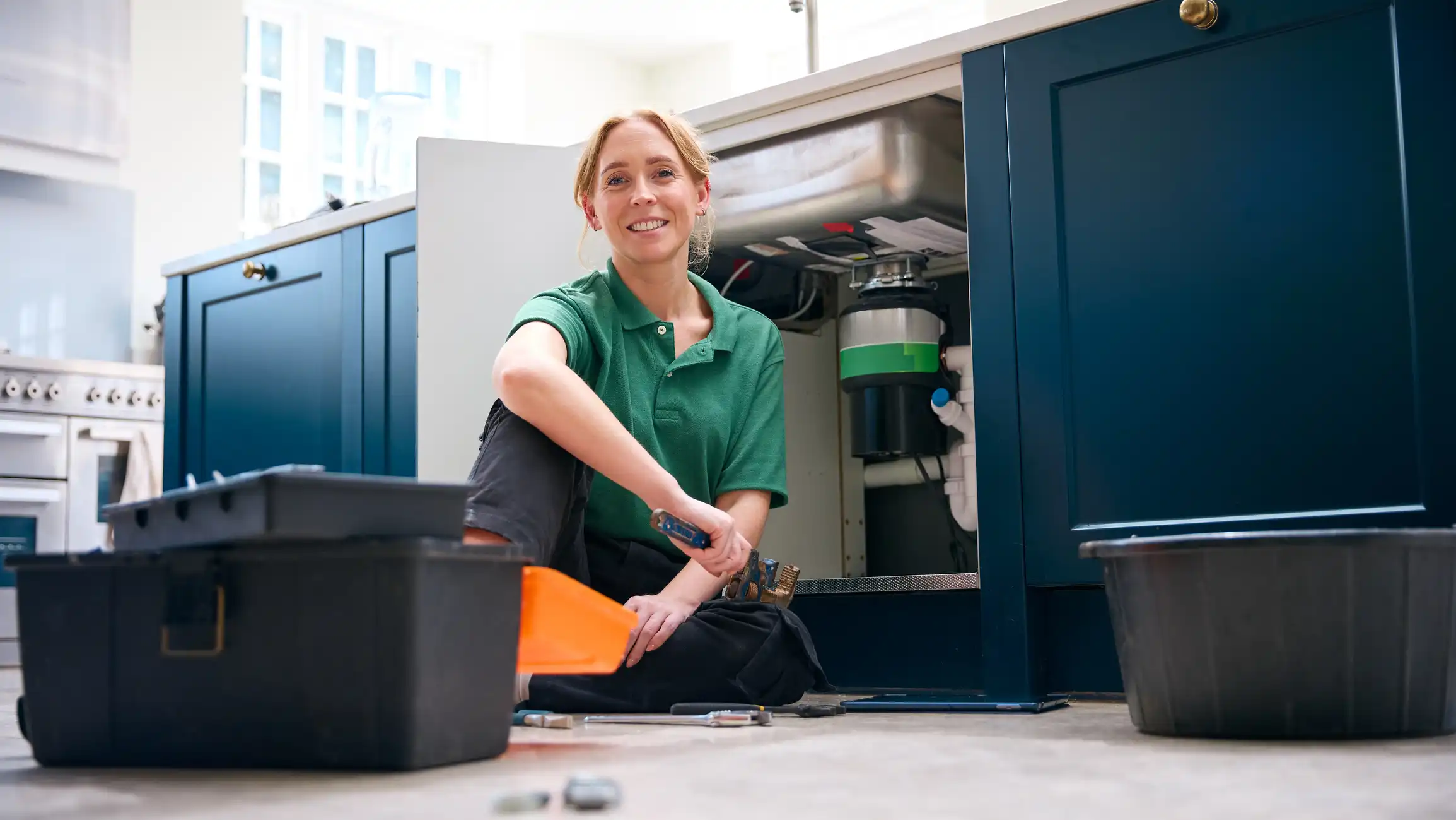 A woman in a green polo shirt sits on the floor, smiling while holding a wrench under a kitchen sink.