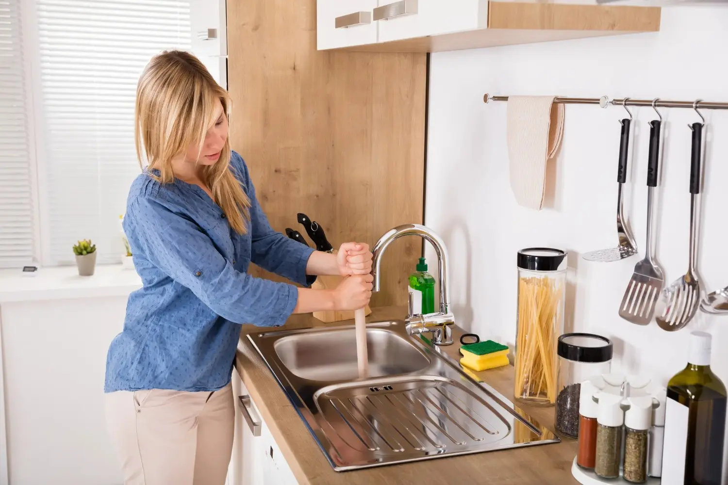 A woman in a blue shirt uses a plunger to unclog a stainless steel kitchen sink.