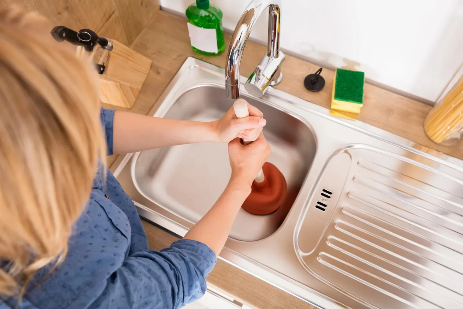 An overhead shot of a woman using a red plunger to unclog a double-basin stainless steel kitchen sink.