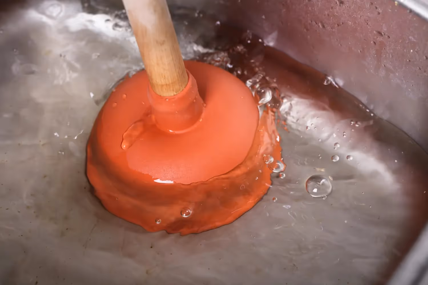 A close-up shot of a red plunger submerged in a stainless steel sink full of water, working to clear a clog.