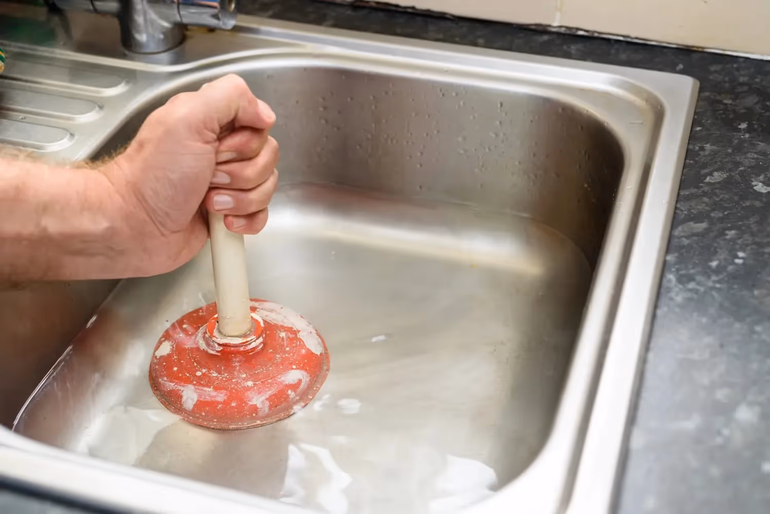 A man's hand uses a red plunger to unclog a stainless steel kitchen sink filled with cloudy water.