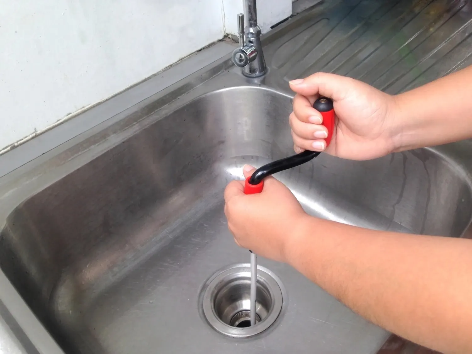 A person's hands use a handheld plumbing snake to clear a clog in a stainless steel sink.