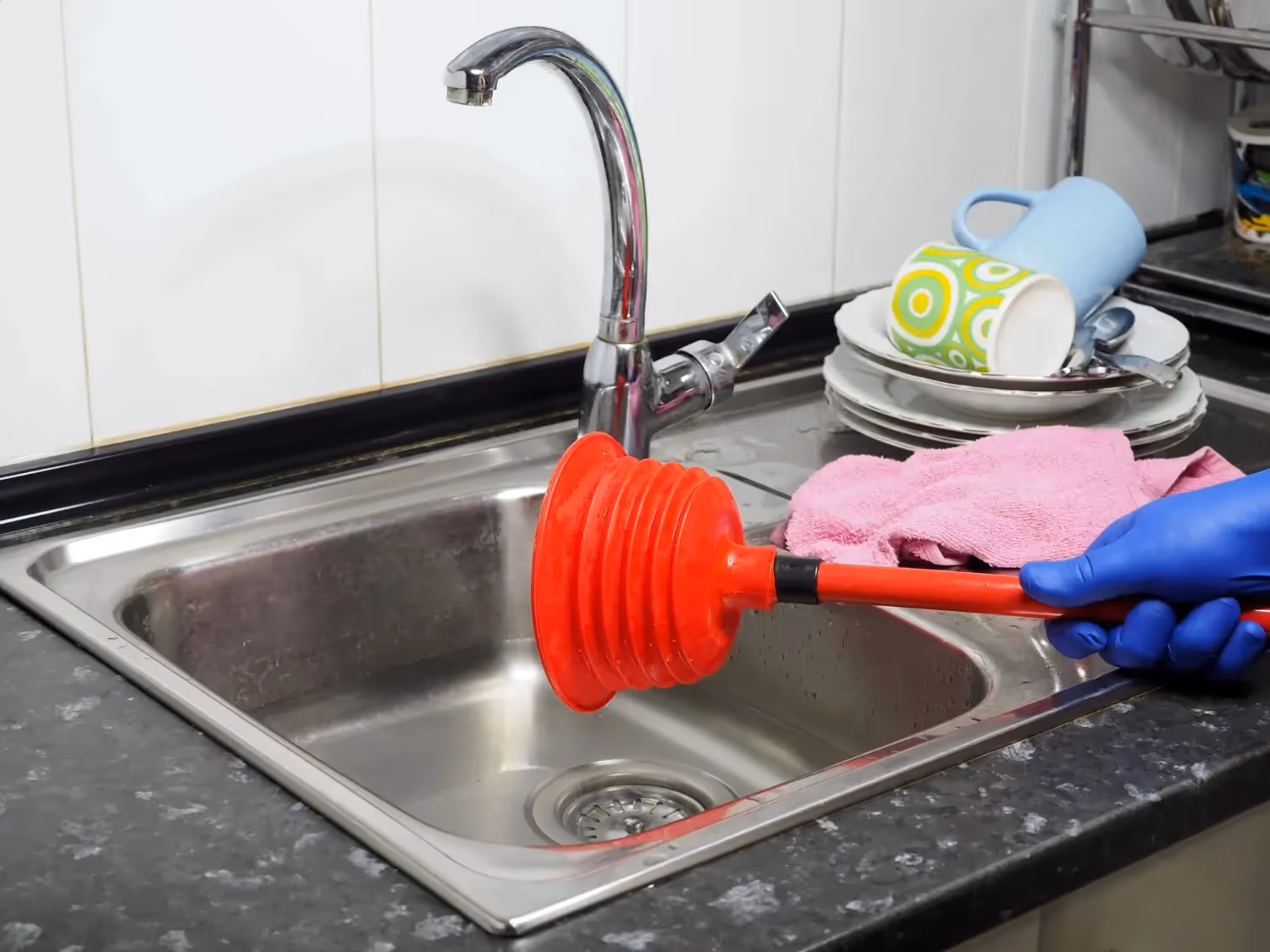 A person wearing blue gloves uses a red plunger in a stainless steel sink.