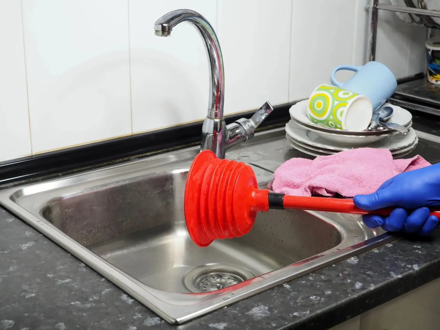 A person wearing blue gloves uses a red plunger in a stainless steel sink.
