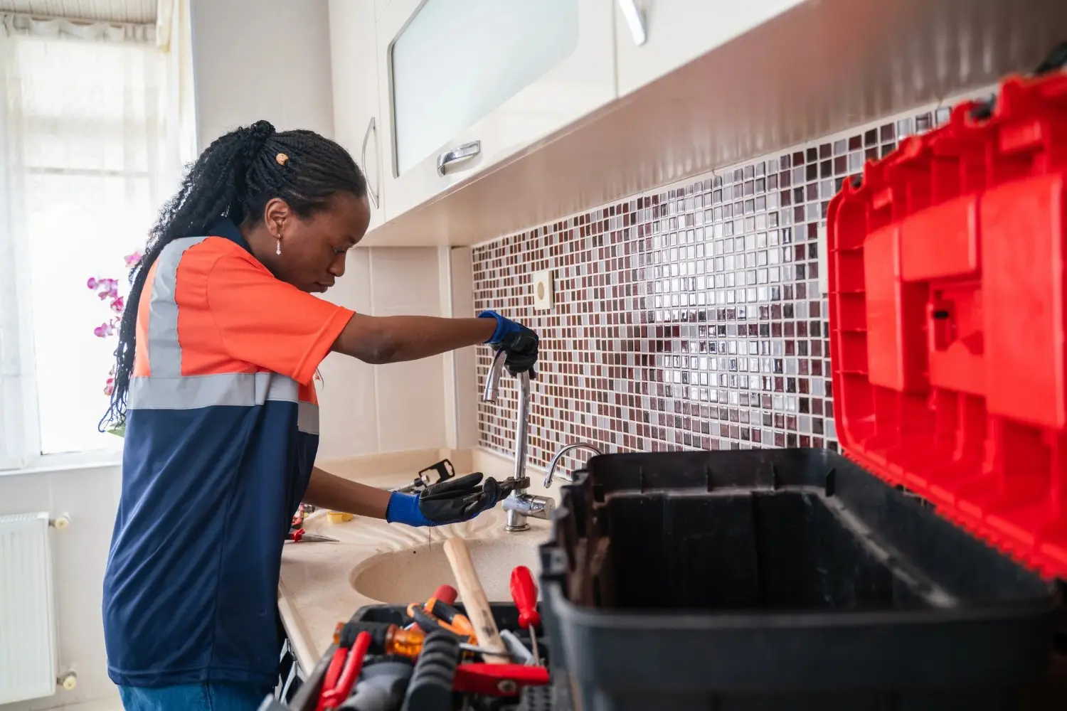A woman in a work uniform and gloves uses a wrench to fix a kitchen sink. 
