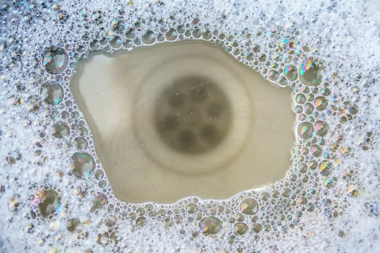 A close-up, high-angle shot shows a drain in a sink surrounded by suds and cloudy water.
