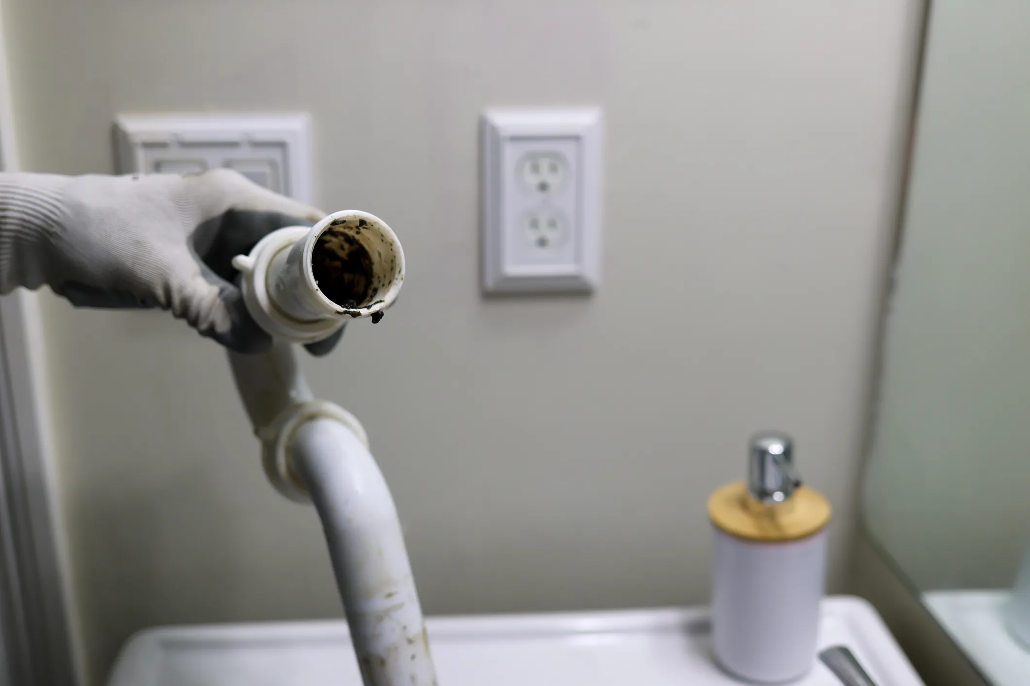 A person's hand wearing a white glove holds a dirty, disconnected pipe from under a sink, with an electrical outlet and a soap dispenser in the background.