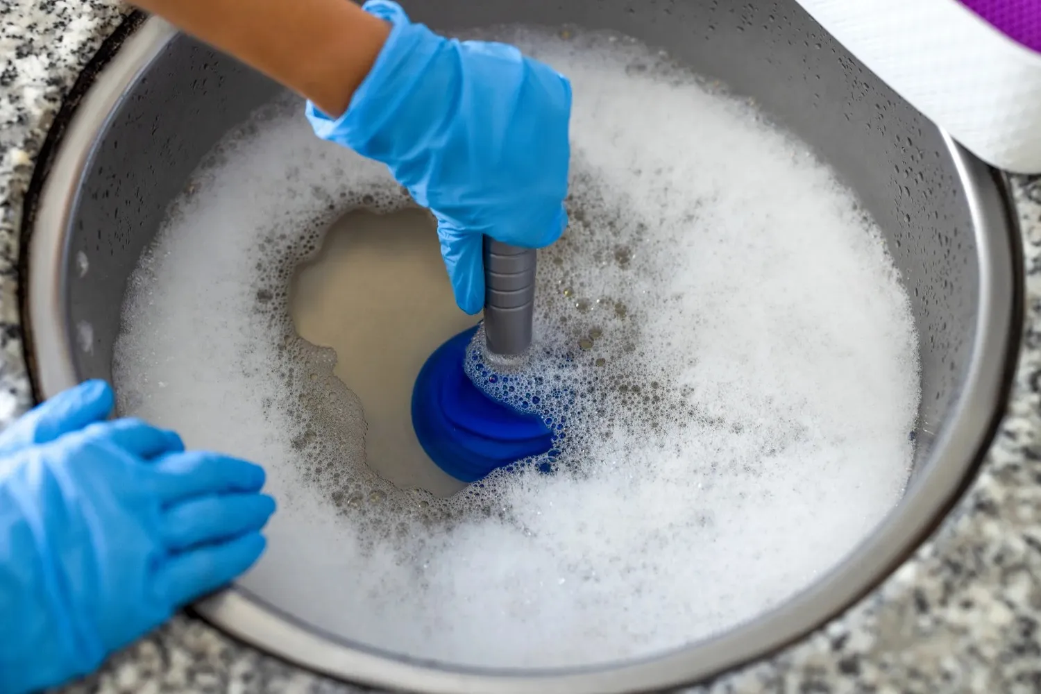 A close-up shot of a person's hands wearing blue rubber gloves, using a blue plunger to clear a clog in a stainless steel sink filled with white soap suds.