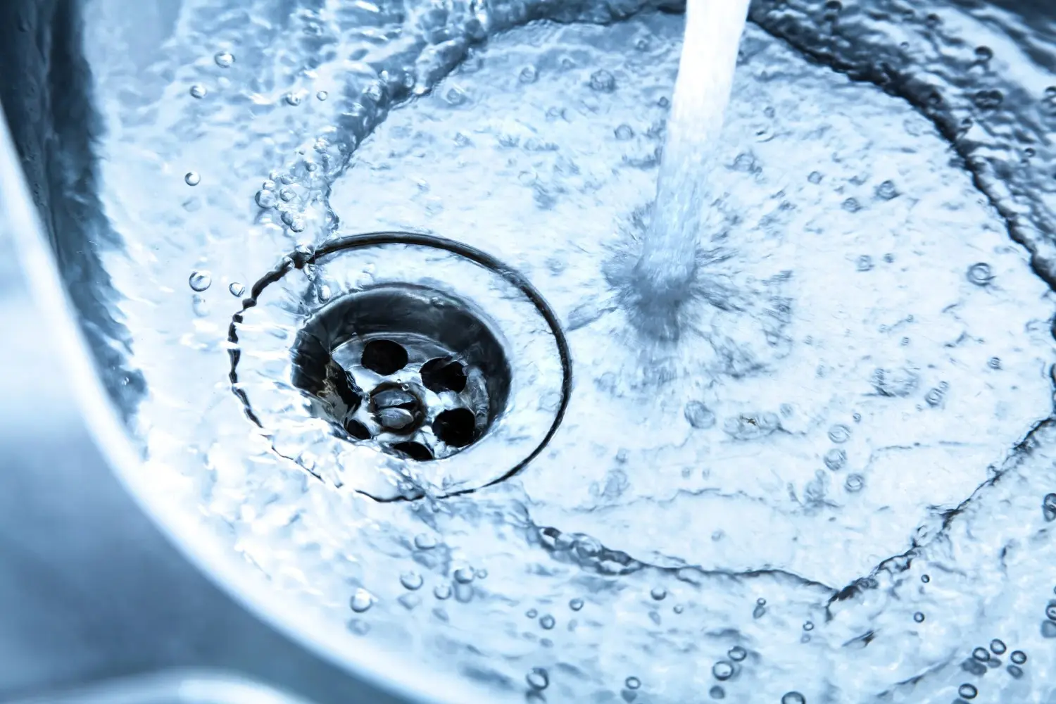 A close-up shot of clear water rushing down into a shiny kitchen sink drain.
