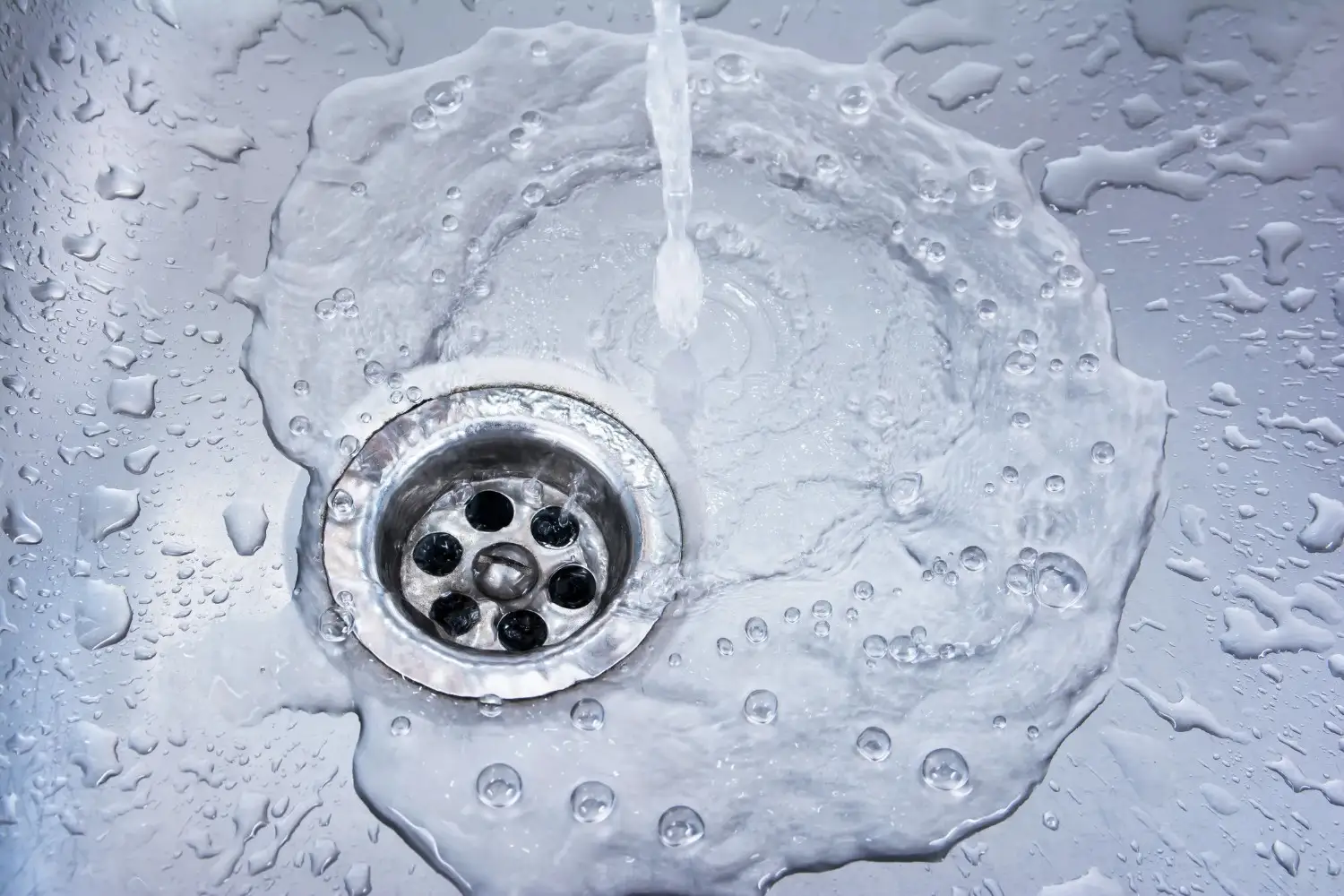 Water running into a stainless steel sink drain, creating a ripple effect on the surface.