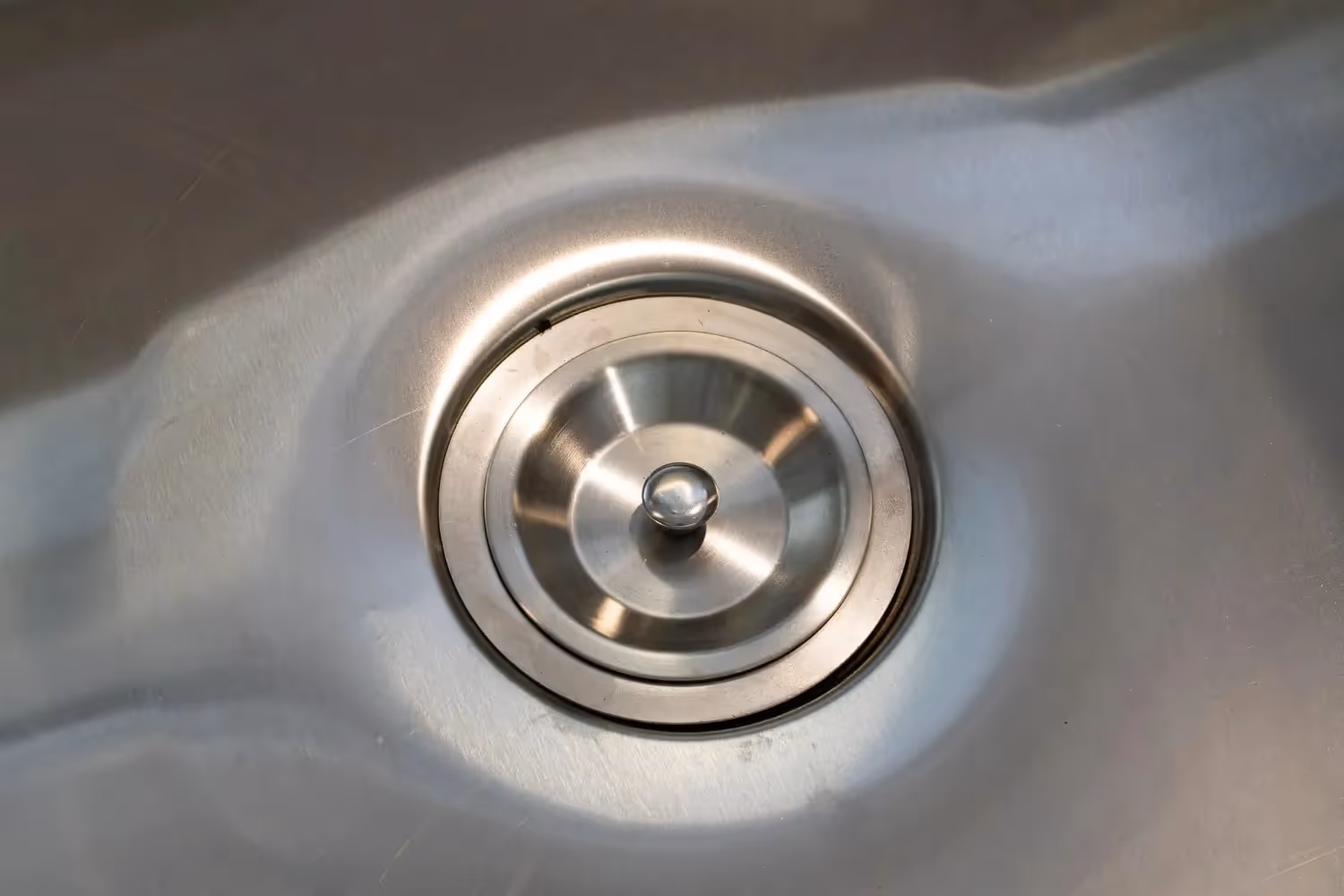 A clean, stainless steel sink drain with the stopper in, reflecting light.