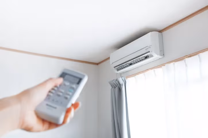 A person's hand holding a remote control pointed at a wall-mounted mini-split air conditioner.