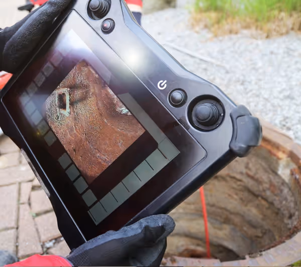 A person holds a tablet displaying sewer camera footage in front of an open hole in the ground.