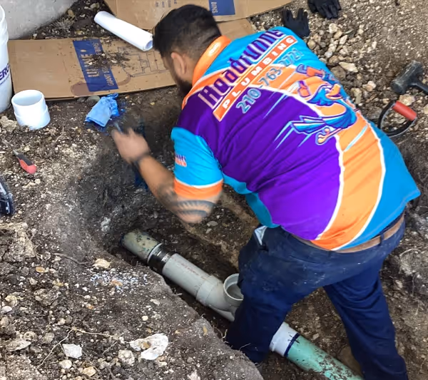 A plumber in a colorful shirt works on underground pipes. He is focused, surrounded by soil, tools, and PVC pipes in a dug-out area.