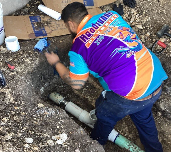 A plumber in a colorful shirt works on underground pipes. He is focused, surrounded by soil, tools, and PVC pipes in a dug-out area.