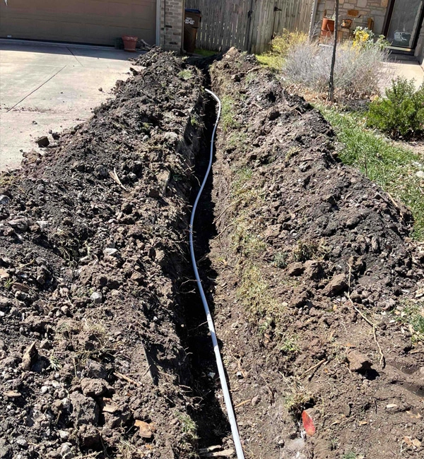 A trench filled with a white pipe runs through brown soil, leading toward a paved driveway in a residential area. The scene suggests recent yard work.