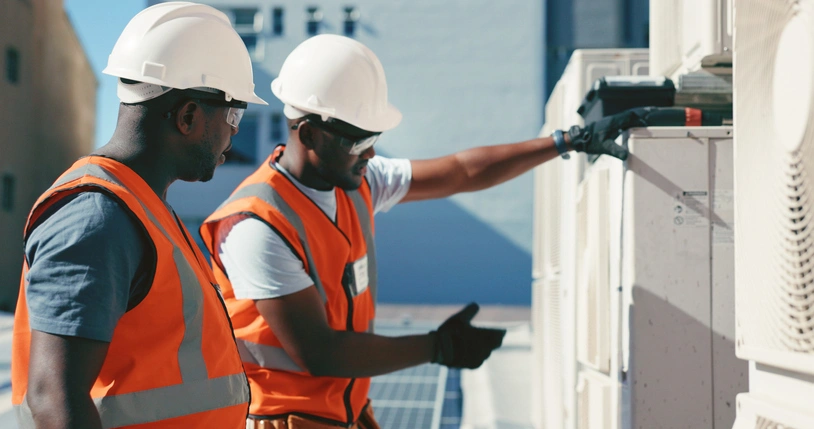 Two workers wearing white hard hats and orange safety vests inspect rooftop HVAC units. 