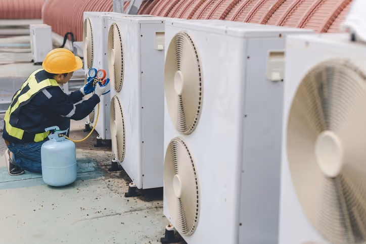 A technician in a yellow hard hat and safety vest inspects rooftop air conditioning units with gauges