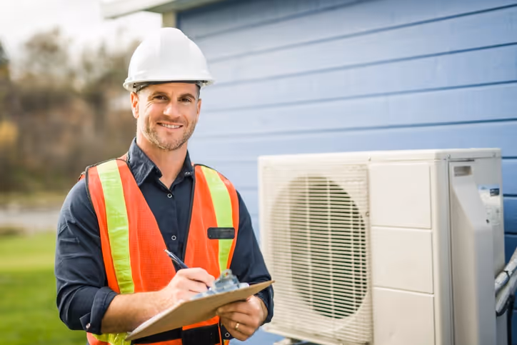 Smiling male construction worker wearing a hard hat and safety vest holds a clipboard near a building with an HVAC unit. The scene conveys professionalism.