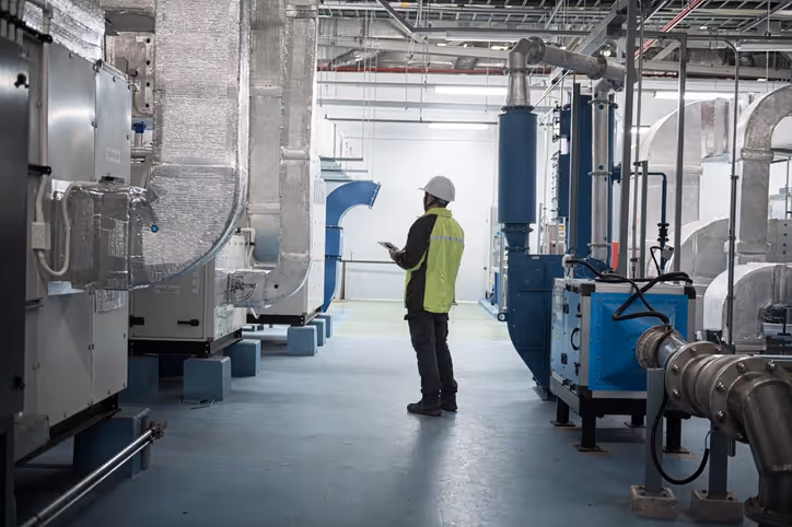 A worker in a hard hat and high-visibility vest inspects industrial machinery in a bright factory. The scene is technical and focused.