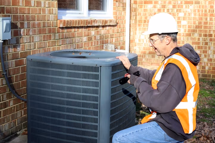 A technician in a hard hat and safety vest inspects an outdoor air conditioning unit beside a brick building