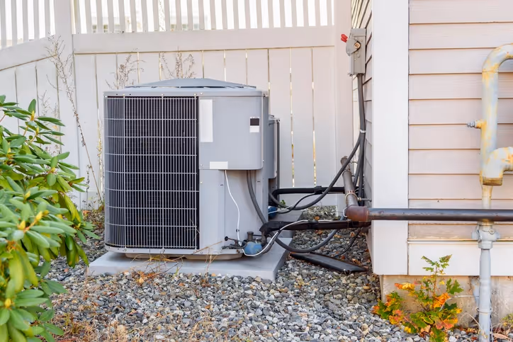 Outdoor air conditioning unit next to a beige house with siding, surrounded by gravel and some greenery. White fence in background, conveying a residential setting.