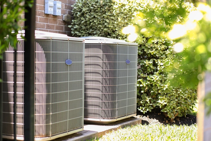 Two air conditioning units are positioned next to a brick wall, surrounded by lush green plants and sunlight