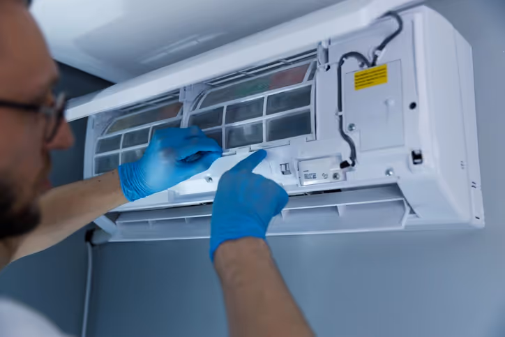 A person wearing blue gloves repairs a wall-mounted air conditioner. They focus on the open front panel, suggesting maintenance or cleaning work.