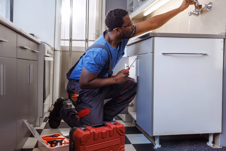 A plumber in a blue shirt and overalls kneels on a checkered floor, fixing a leaking sink. 