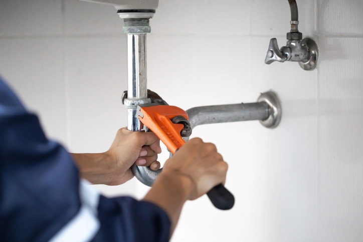 A person in a blue uniform uses an orange wrench to tighten a pipe under a sink. 