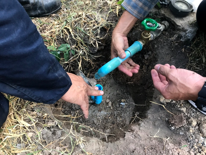 Plumbers work together to repair a water pipe in a dirt trench. Hands hold blue pipes and a green valve.