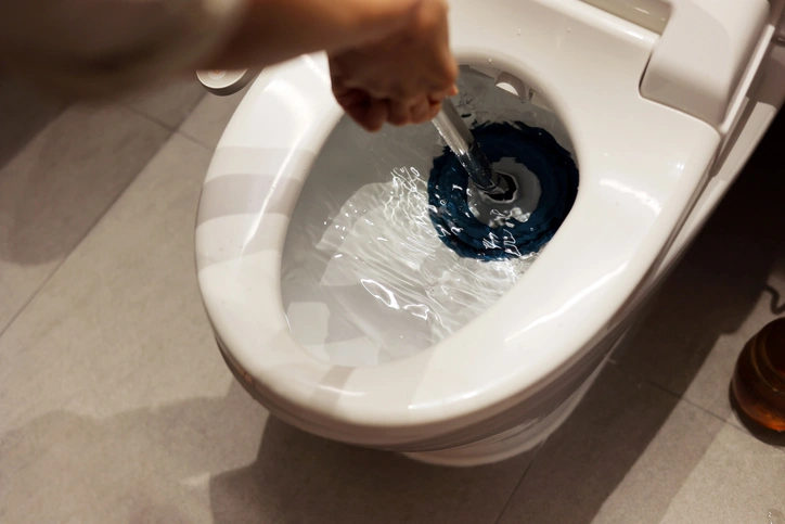 A person is cleaning a white toilet with a brush, scrubbing blue cleaner in the bowl. The setting is a bathroom with a tiled floor. The mood is practical and focused.