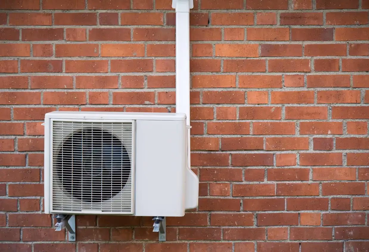 A white, wall-mounted heat pump unit is installed on a red brick wall, with a white pipe running up the side.