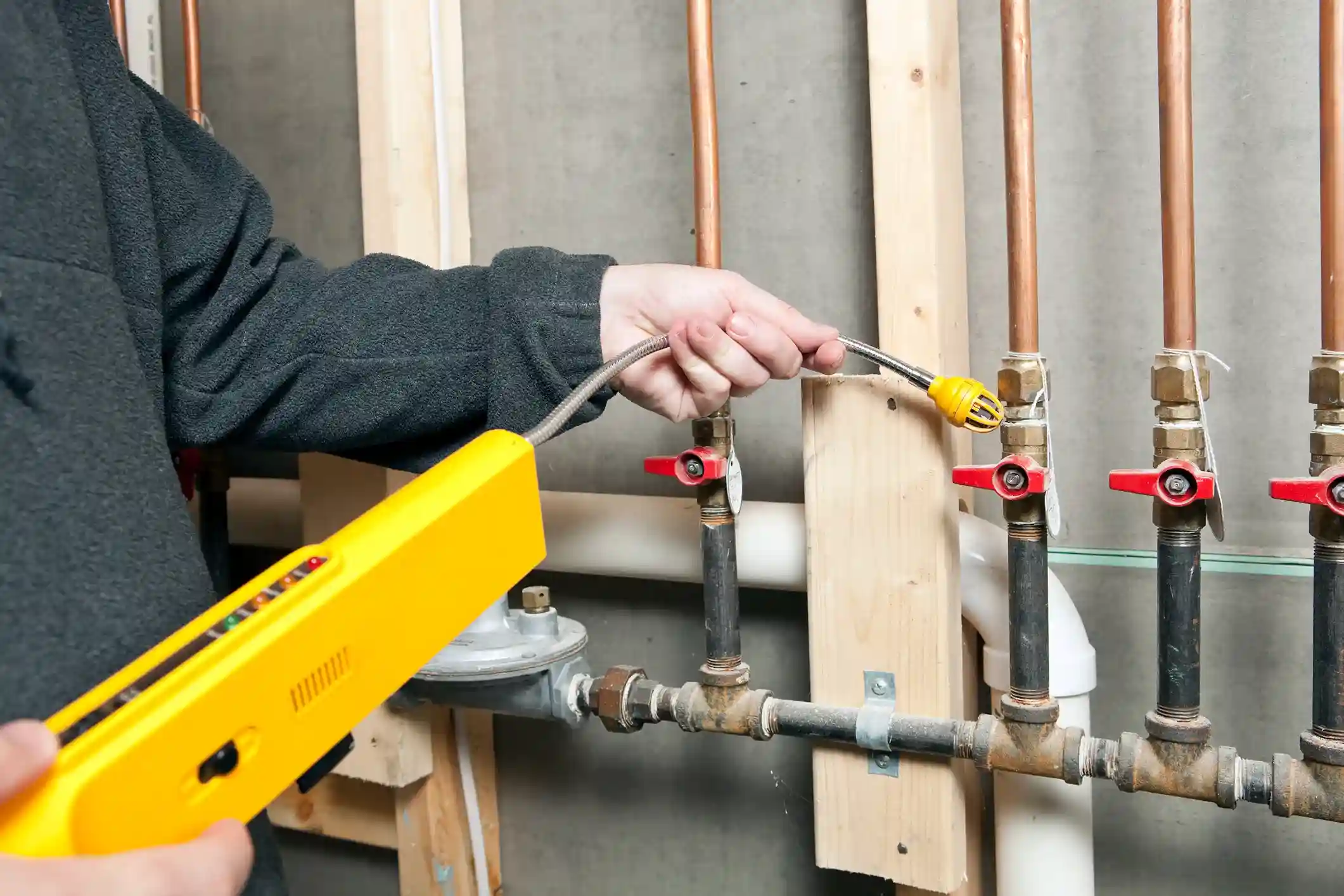 A person is using a yellow gas leak detector with a flexible probe to check for leaks on a series of copper pipes and red valves.