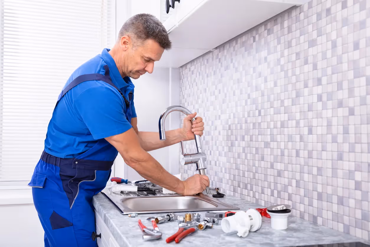 A plumber in blue overalls is working to install a new chrome faucet on a kitchen sink with a gray tile backsplash.