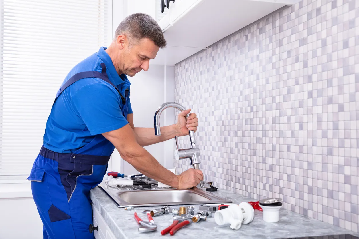 A plumber in blue overalls is working to install a new chrome faucet on a kitchen sink with a gray tile backsplash.