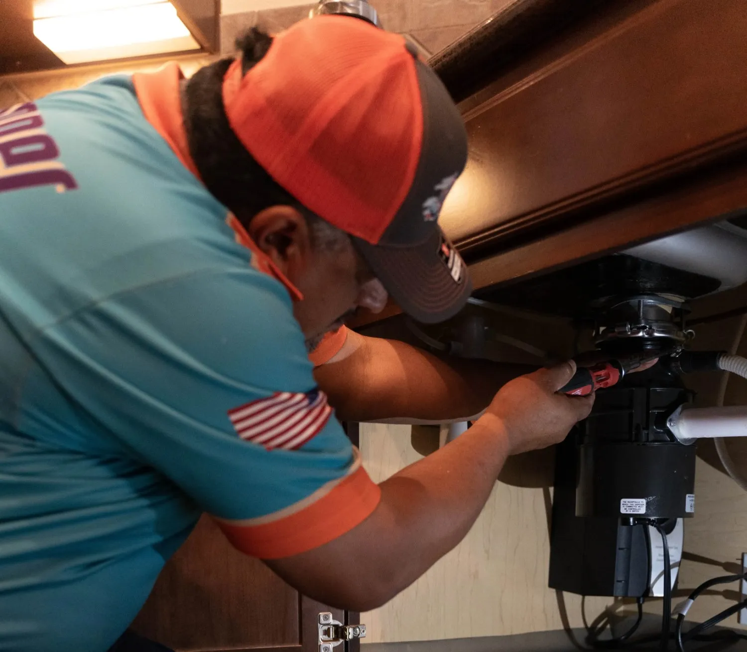 A plumber wearing an orange hat repairs a kitchen garbage disposal under a sink