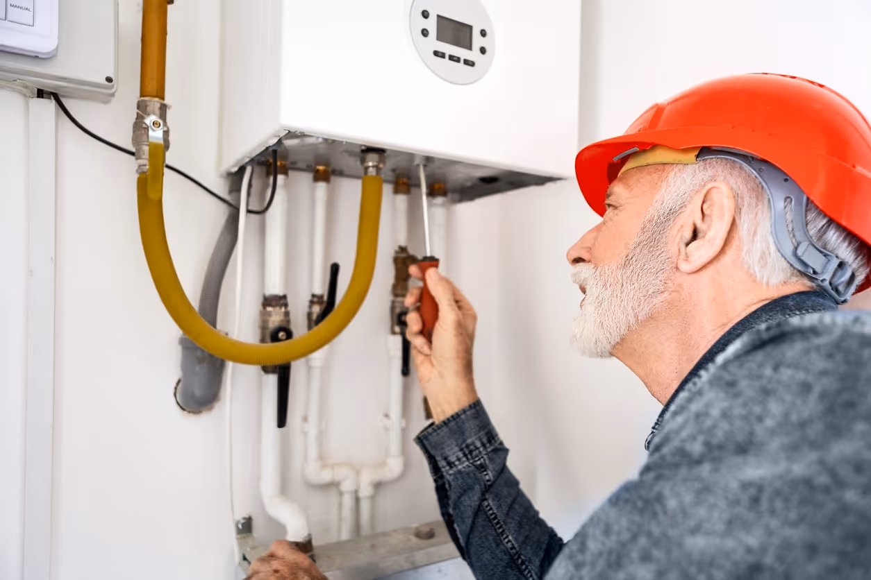 Elderly man in a red hard hat uses a screwdriver to fix a wall-mounted boiler. 