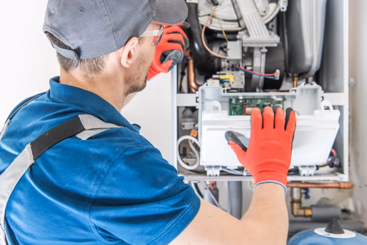 A furnace technician wearing a blue shirt cap and red gloves working on the internal components of a furnace