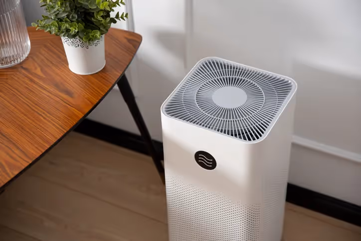 A square, white air purifier with a circular grille on top sits on a wood floor next to a modern side table with a potted plant.