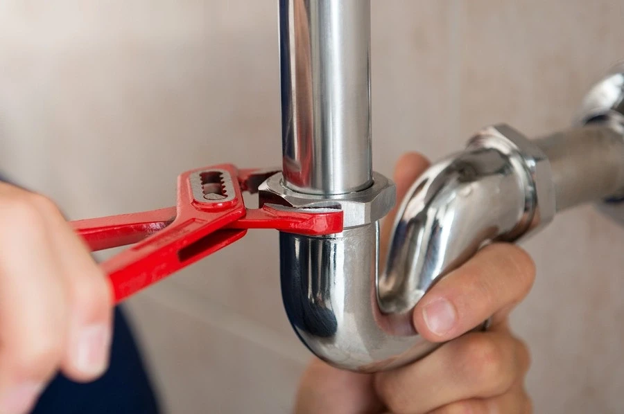 A red adjustable wrench is being used to tighten a nut on a chrome P-trap pipe under a sink.