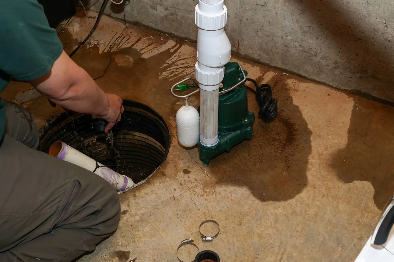 A man is crouched on the floor, working on an open sump pump pit and a green pump unit with white PVC piping.