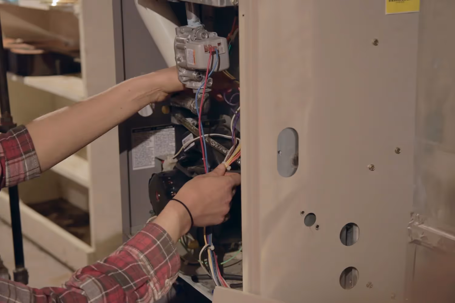  A technician wearing a red and white plaid shirt is performing a repair or inspection on the internal components of a gas furnace. The side panel of the furnace is removed, and the technician's hands are visible as they work with a bundle of colored electrical wires connected to the gas valve assembly and other parts inside the unit.