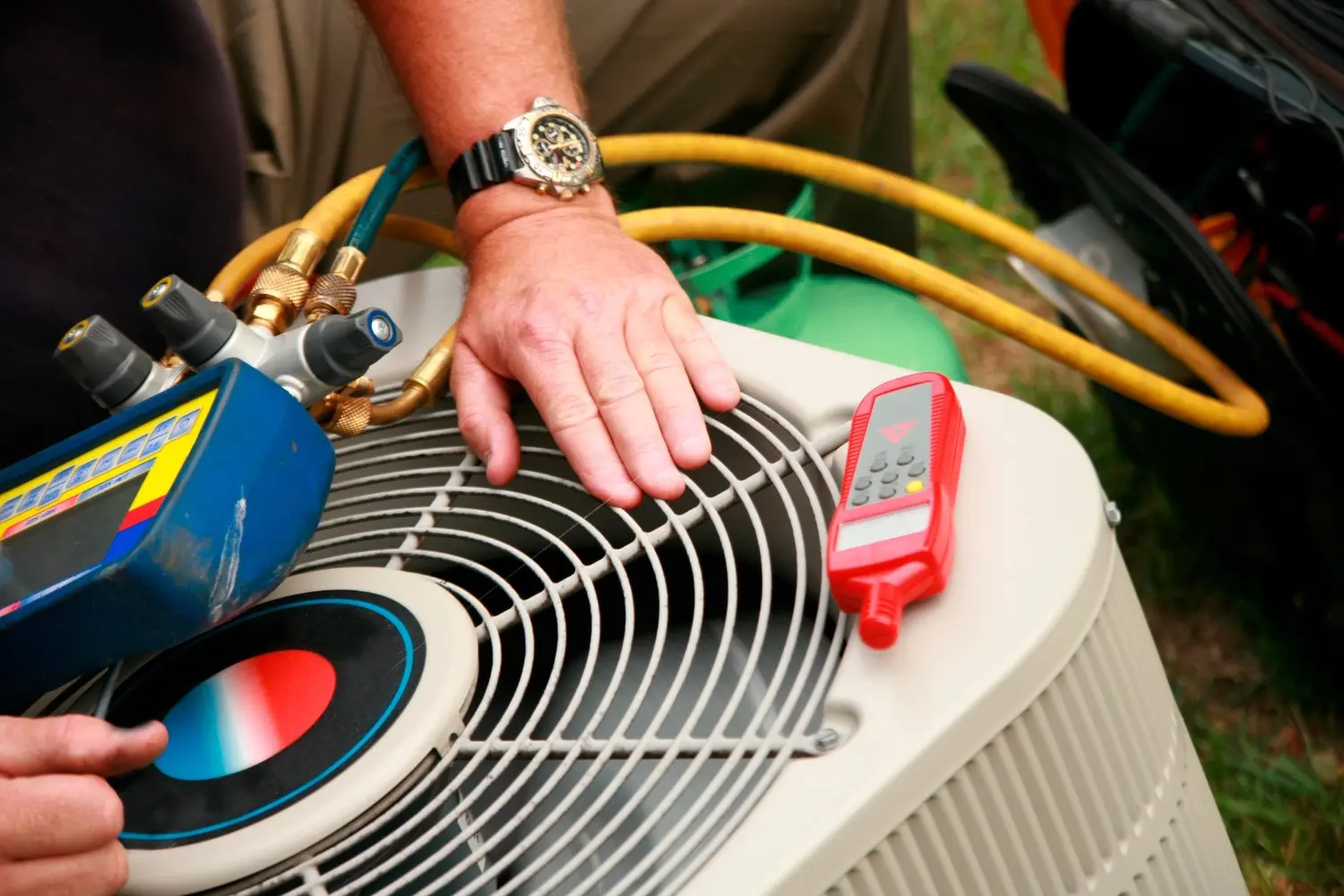 A technician's hands and tools, including a manifold gauge and a red remote, on the fan grille of an outdoor AC condenser unit.