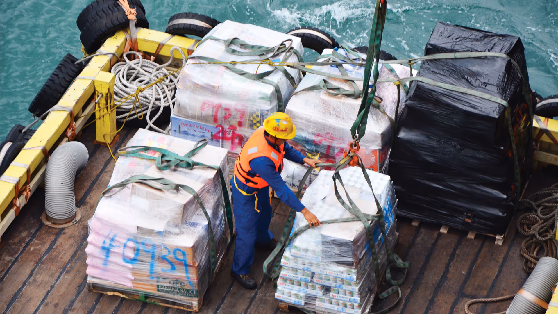 Seafarer in safety vest and helmet securing cargo wrapped in plastic on a ship deck by the sea.