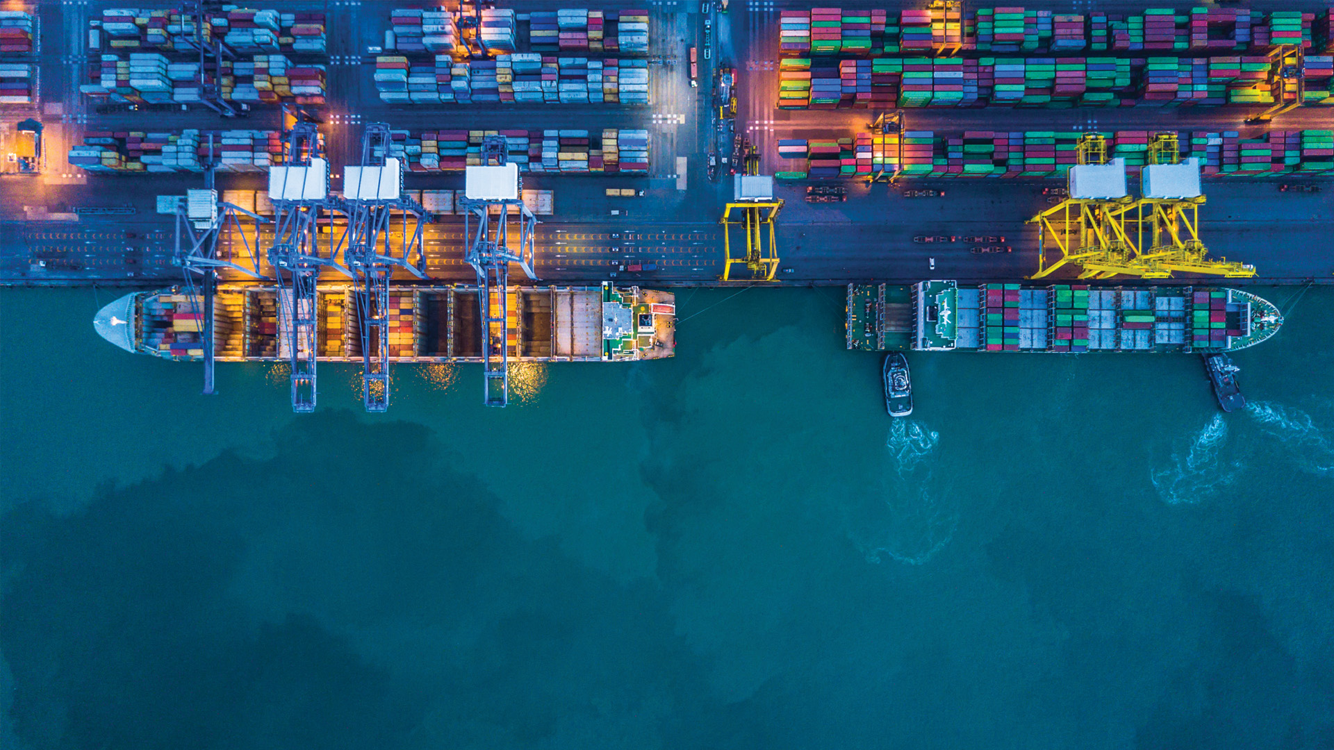 Aerial view of two cargo ships docked at a port with colorful shipping containers and cranes.