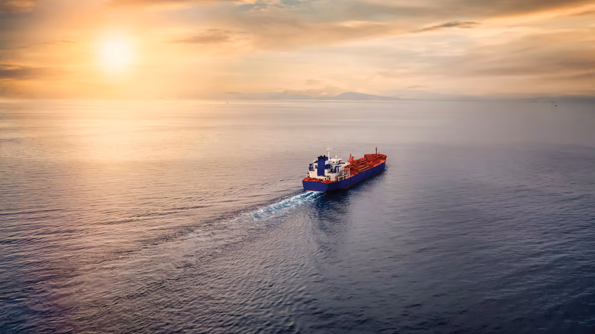 Red and blue cargo ship sailing on calm ocean waters at sunset with a distant mountain horizon.